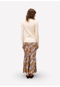 Femme aux cheveux bruns mi-longs portant un pull côtelé crème et une longue jupe marron à motifs, se tenant de dos sur fond blanc.