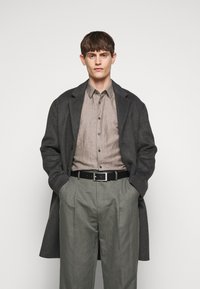 Grijs wollen overjas over een lichtgrijze shirt, met grote geplooide grijze broeken. Zwarte riem met een rechthoekige zilveren gesp.