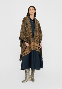 Šál s leopardím vzorom v hnedej a čiernej farbe, s třásňami na okrajoch, nosený cez tmavé džíny šaty, v kombinácii s lesklými striebornými topánkami s hadím vzorom.