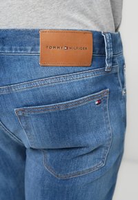 Μπλε τζιν παντελόνι από ντένιμ με καφέ δερμάτινο ταμπελάκι που φέρει ανάγλυφο το λογότυπο "TOMMY HILFIGER" και κόκκινες, λευκές και μπλε λεπτομέρειες στην πίσω τσέπη.