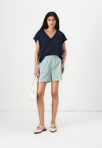 Haut marin à manches courtes avec décolleté en V, short texturé vert clair, chaussures beiges avec des lanières discrètes, et un petit sac à main blanc.