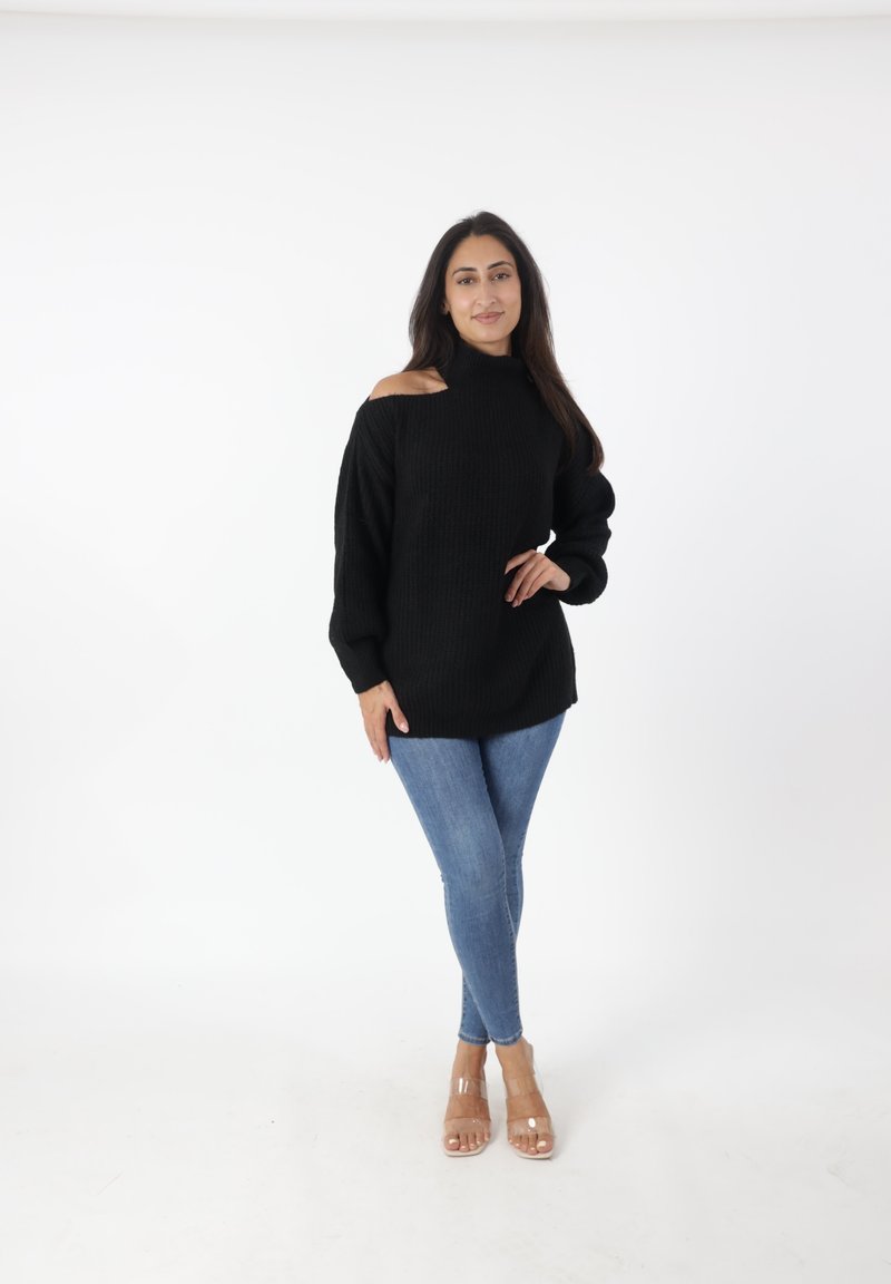 Maglione nero lavorato a maglia con design a spalle scoperte, maniche lunghe e tessuto testurizzato, abbinato a jeans blu e sandali con tacco trasparente.
