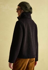 Czarna polarowa kurtka z wysokim kołnierzem, teksturowaną powierzchnią i subtelnym logo z tyłu. Połączona z brązowymi spodniami z sztruksu.