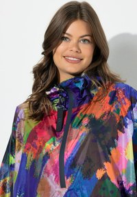 Giacca impermeabile leggera multicolore con cappuccio con coulisse, chiusura frontale con cerniera e motivi astratti che richiamano pennellate in tonalità vivaci.