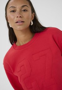 Czerwone bluzy mają wytłoczone wzory '27', okrągły dekolt i gładką teksturę. Modelka nosi srebrne kolczyki w kształcie kół.