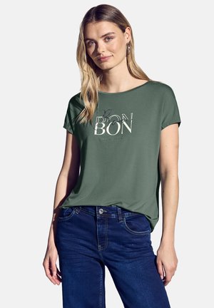 Grøn kortærmet t-shirt med et trykt design, der har ordet "BON" i forskellige skrifttyper. Kombineret med mørkeblå jeans.