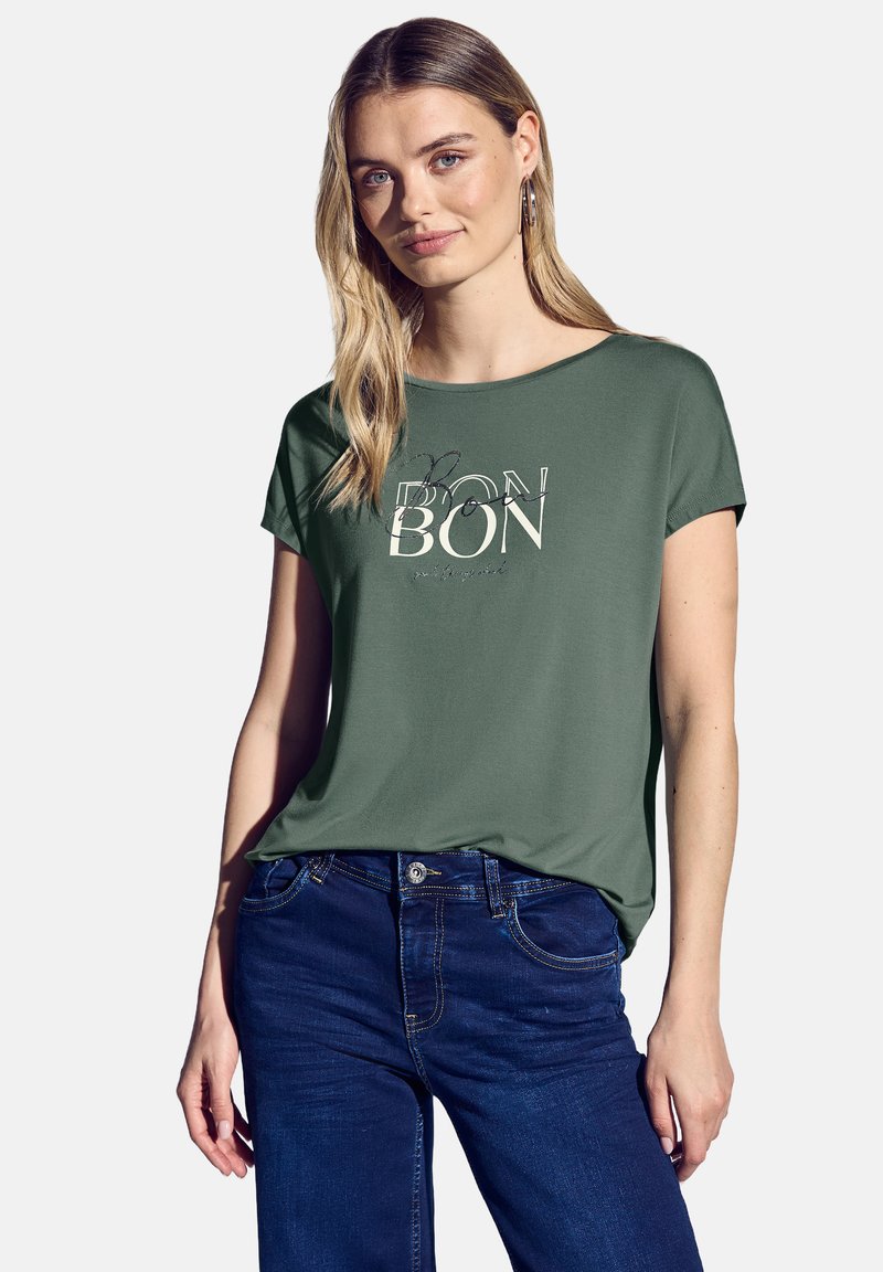 Zelené tričko s krátkým rukávem s tištěným designem obsahujícím slovo "BON" v různých písmových stylech. Spárované s tmavě modrými džíny.