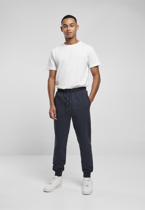 Jaunas vyras stovintis viena ranka kišenėje, dėvintis baltą marškinėlį, tamsiai mėlynas stringuotas sportines kelnes ir baltus sportbačius, priešais paprastą foną.