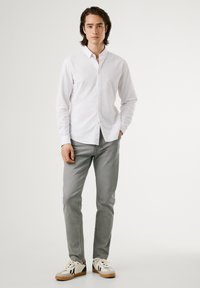 Junger Mann, der ein weißes Hemd mit Knöpfen, hellgraue Jeans und weiße Sneakers trägt, steht vor einem schlichten weißen Hintergrund.