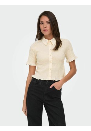 Femme aux longs cheveux foncés portant un haut crème à manches courtes boutonné avec des bords volantés et un pantalon noir taille haute, debout avec une main dans la poche.