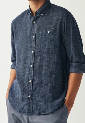 Hombre vistiendo una camisa azul marino texturizada abotonada, con las mangas arremangadas y las manos en los bolsillos, combinada con pantalones grises.