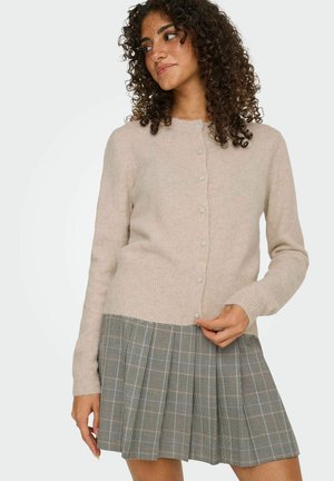 Cardigan beige clair avec un col rond, des manches longues et des boutons nacrés, porté par-dessus une jupe plissée en tartan gris.