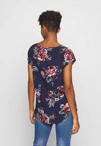 Top floreale navy a maniche corte, con rose rosa e bianche, scollatura rotonda, piccola apertura a chiave sul retro e orlo curvo. Abbinato a jeans blu.