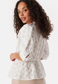 Witte, textuurgegeven blouse met een peplumfit, driekwart mouwen en een subtiel ruitpatroon, ontworpen met een ronde halslijn.