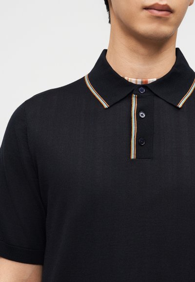 Άνδρας που φοράει μαύρο πόλο μπλουζάκι με κοντά μανίκια και πολύχρωμη ριγέ επένδυση στο γιακά και στη λωρίδα με τα κουμπιά.