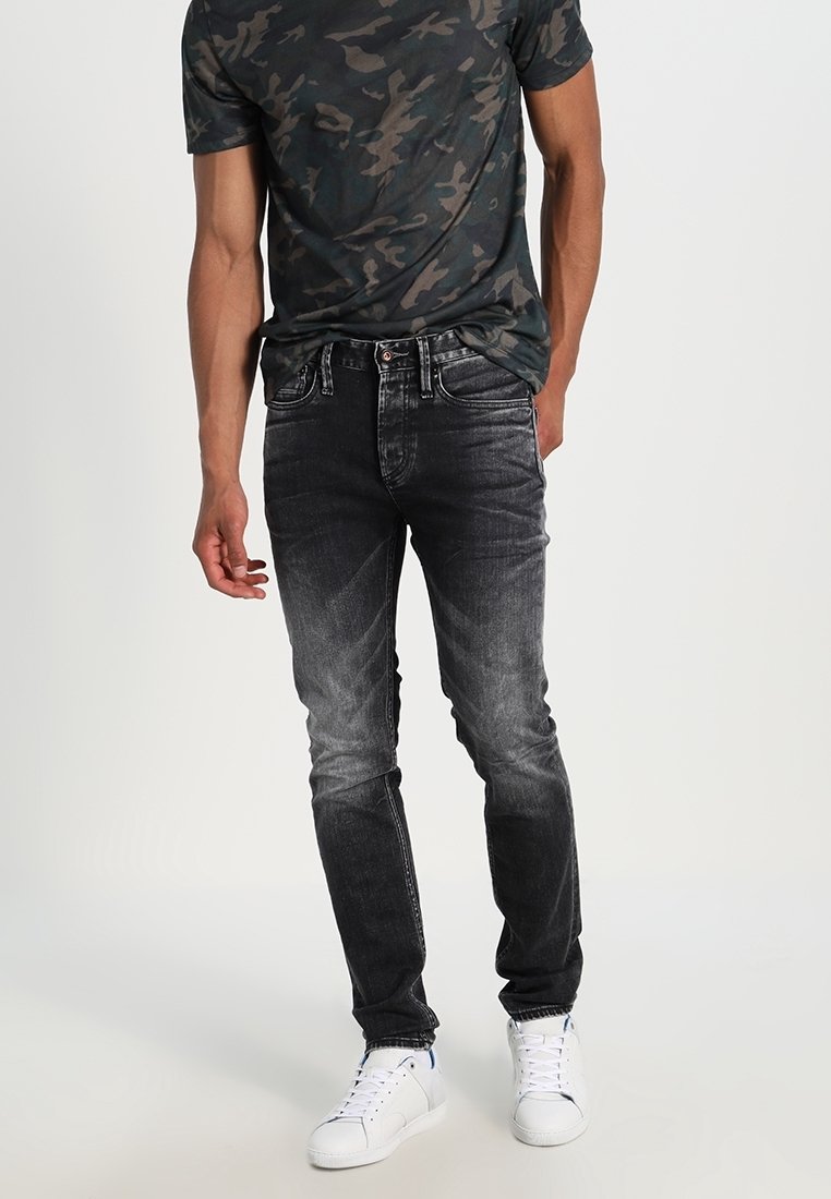 Homme portant un t-shirt camouflage foncé, un jean skinny noir délavé et des baskets blanches, debout devant un fond clair et uni.