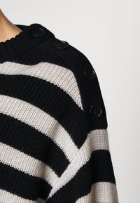 Sweter dziergany z poziomymi paskami w kolorze czarnym i kremowym, z textured'owym splotem i dużymi czarnymi guzikami na ramieniu.