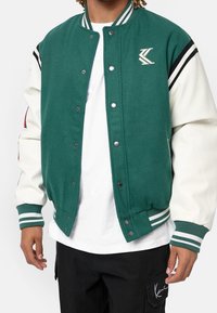 Giacca varsity verde con maniche color crema, polsini e colletto a costine. Presenta strisce bianche e dettagli del logo sul petto. Chiusura a bottoni.