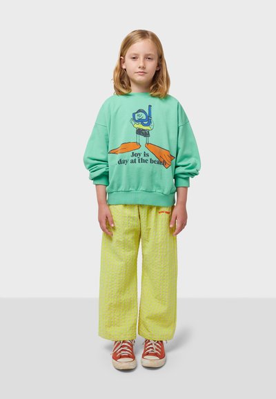 Felpa oversize verde menta con grafica da spiaggia, abbinata a pantaloni a gamba larga giallo brillante e testurizzati e sneakers marroni.