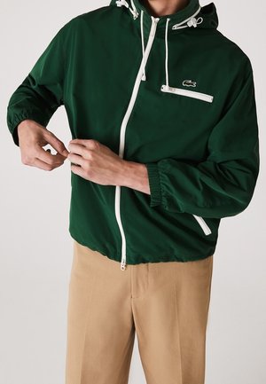 Personne attachant une veste zippée vert foncé avec des fermetures éclair blanches et des cordons de serrage, portant un pantalon beige, visible du cou jusqu'au milieu des cuisses.