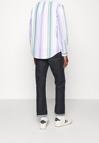Camicia a righe in colori pastello, abbinata a jeans in denim scuro e sneakers bianche con dettagli verdi. Maniche arrotolate visibili.
