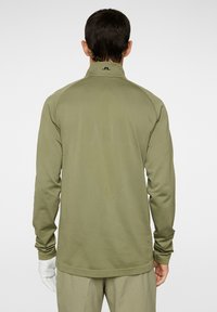 Olivgrönt långärmad topp med hög krage, strukturerat mönster och kontrasterande svart logotyp på baksidan. Lätt vikt, slim fit design.