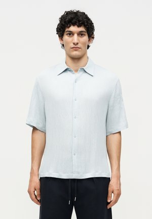 Άνδρας με σκούρα, σγουρά μαλλιά που φοράει ανοιχτό μπλε κοντομάνικο πουκάμισο με κουμπιά και μαύρο παντελόνι, στέκεται μπροστά σε ένα απλό ανοιχτό φόντο.