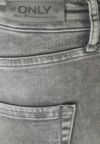 Jean cinza em denim com um acabamento texturizado, detalhes de costura, e um rótulo que diz "ONLY Blue Denim anno 1995." Costura de destaque visível.