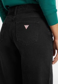 Jeans in denim nero con un classico design a cinque tasche, caratterizzati da un patch triangolare rosso e bianco sulla tasca posteriore e tessuto testurizzato.
