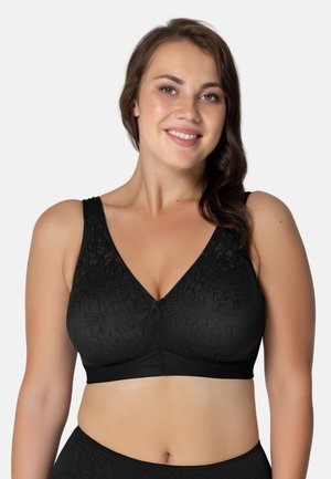 Uśmiechnięta kobieta w czarnym biustonoszu z wzorem i dopasowanej bieliźnie, zwrócona przodem na jednolitym jasnym tle.