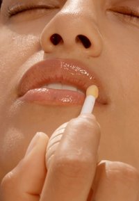 Un lucidalabbra di un leggero colore beige viene applicato con un piccolo applicatore rotondo. Le labbra sono lucide, riflettendo la luce, con la texture naturale visibile.
