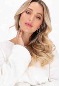 Weißer Pullover mit weiten Ärmeln, welliges blondes Haar, silberne Creolen und neutrale Make-up mit sanften rosanen Lippen und warmen Lidschatten.