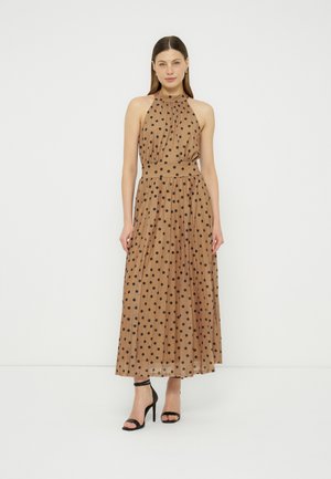 Γυναίκα στέκεται φορώντας ένα αμάνικο μπεζ μαξι φόρεμα με μαύρα πουά και μαύρα σανδάλια με ψηλό τακούνι σε λευκό φόντο.