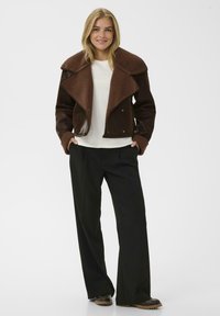 Giacca in pelle marrone con un ampio colletto in pile marrone, abbinata a un top bianco e pantaloni neri a gamba larga. Design a taglio dritto, hardware minimale.