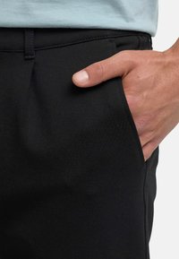 Høyre hånd hviler inne i frontlommen på svarte bukser, båret med en lys skjorte som er stukket inn.