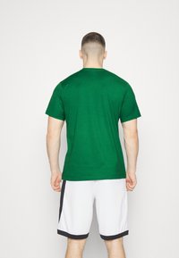 Mies seisoo selkä kameraan päin, päällä vihreä lyhythihainen paita ja valkoiset urheilushortsit mustilla yksityiskohdilla, taustalla yksinkertainen tausta.