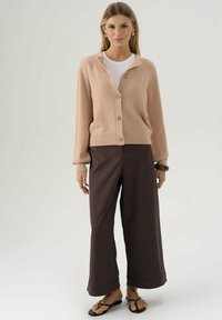 Donna che indossa un cardigan beige con bottoni sopra una camicia bianca, pantaloni larghi neri e sandali neri, con orecchini a cerchio, in piedi contro uno sfondo neutro.