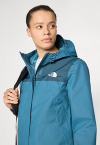 Teal waterproof jacket with a contrasting dark blue upper section, zip front, adjustable hood, and logo detail. Lightweight, smooth texture.