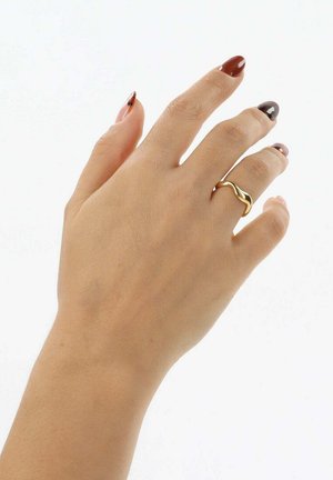 Anillo de diseño en forma de ola dorada sobre una mano de tono de piel claro, acentuado con esmalte de uñas oscuro y neutro en las puntas de los dedos. Textura suave, forma elegante.