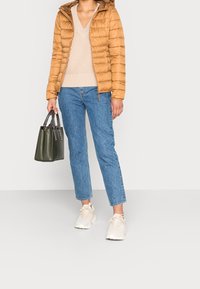 Beige Pufferjacke, cremefarbener, gestrickter V-Ausschnitt-Pullover, blaue gerade Jeans und weiße Sneakers. Grünes Handtaschen mit doppelten Henkeln.