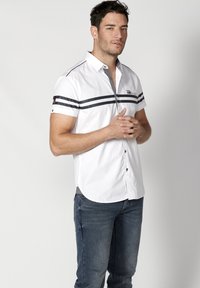 Camisa blanca de manga corta con franjas horizontales oscuras en el pecho, cuello puntiagudo y cierre de botones en el frente, combinada con jeans azules.