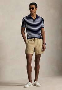 Άντρας με γυαλιά ηλίου, μπλε ναυτικό και λευκό ριγέ πόλο, μπεζ σόρτς με κορδόνι, λευκά αθλητικά παπούτσια, στέκεται με τα χέρια στις τσέπες.