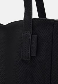 Czarna torba tote z teksturowaną powierzchnią, solidnymi uchwytami i małą metką z logo, prezentująca elegancki, minimalistyczny design.