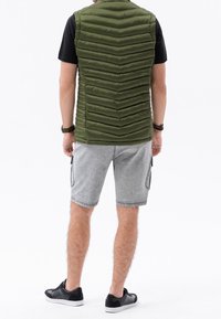 Mężczyzna ubrany w oliwkowozieloną pikowaną kamizelkę, czarną koszulkę, szare szorty typu cargo, czarne sportowe buty i opaski na nadgarstki, stojący tyłem na białym tle.