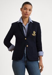 Blazer azul-marinho com botões dourados e detalhe de bolso bordado, combinado com uma camisa às riscas azul e branca com punhos dobrados.