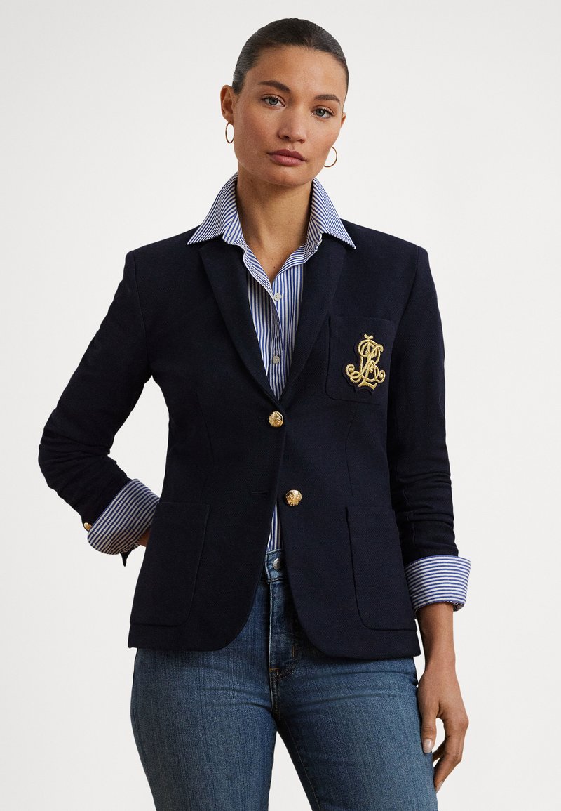 Blazer azul-marinho com botões dourados e detalhe de bolso bordado, combinado com uma camisa às riscas azul e branca com punhos dobrados.