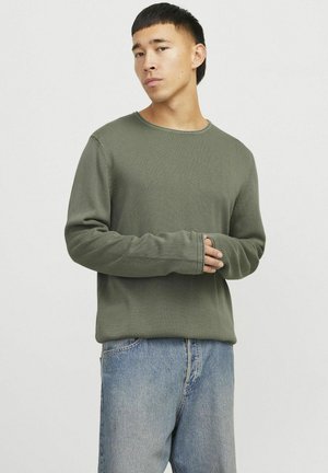 Olivgrünes Langarmshirt mit rundem Ausschnitt, gerippter Textur und verlängerten Manschetten, kombiniert mit hellblauen Jeans.
