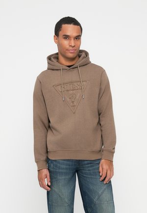 Hombre vistiendo una sudadera marrón de Guess con un logo frontal y jeans azules, de pie contra un fondo blanco liso.
