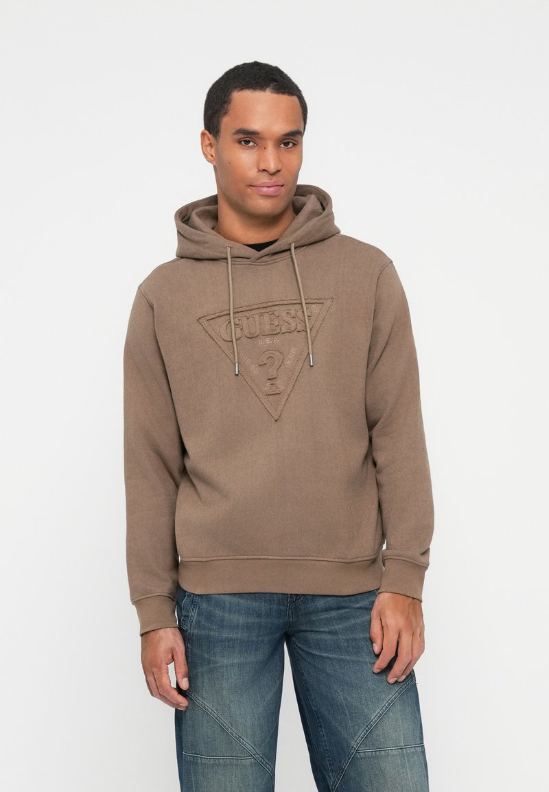 Hombre vistiendo una sudadera marrón de Guess con un logo frontal y jeans azules, de pie contra un fondo blanco liso.