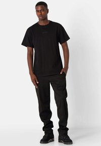 Zwarte katoenen T-shirt met korte mouwen en een ronde hals, met subtiele geperste tekst op de voorkant. Gecombineerd met zwarte broek en sneakers.